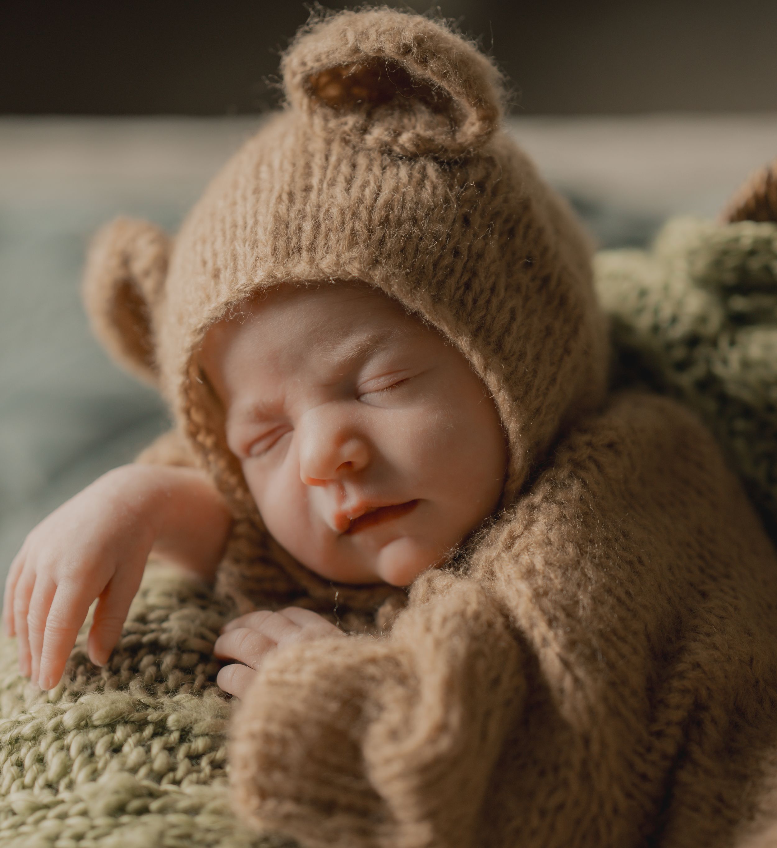 Sleeping baby wearing cozy brown knitted bear hat resting on soft green blanket.