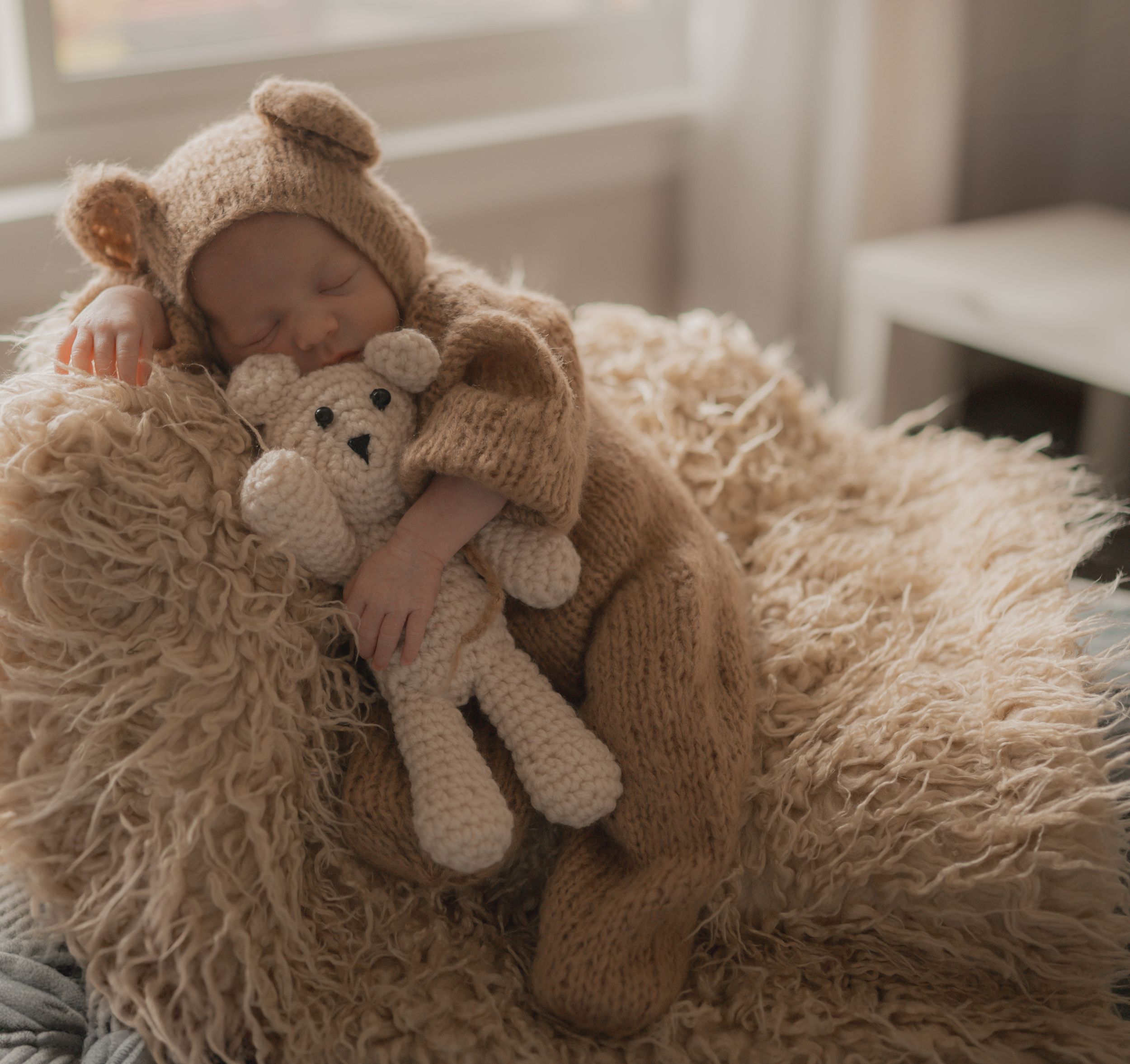 A sleeping baby in cozy beige knitwear lies on fluffy carpet holding a knitted teddy bear toy.