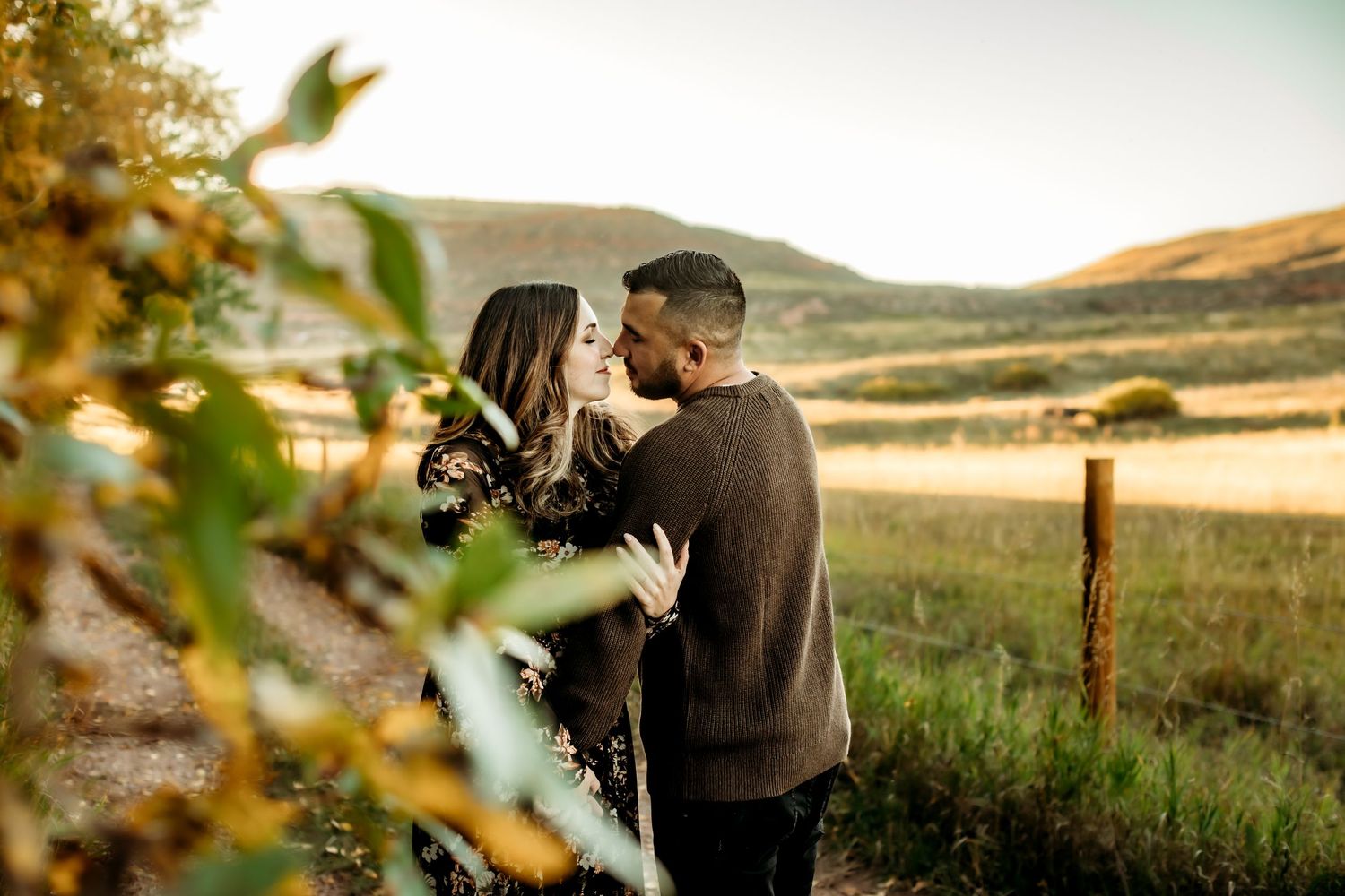 A couple in dark clothing embraces on a dirt path during golden hour with autumn foliage in the background.