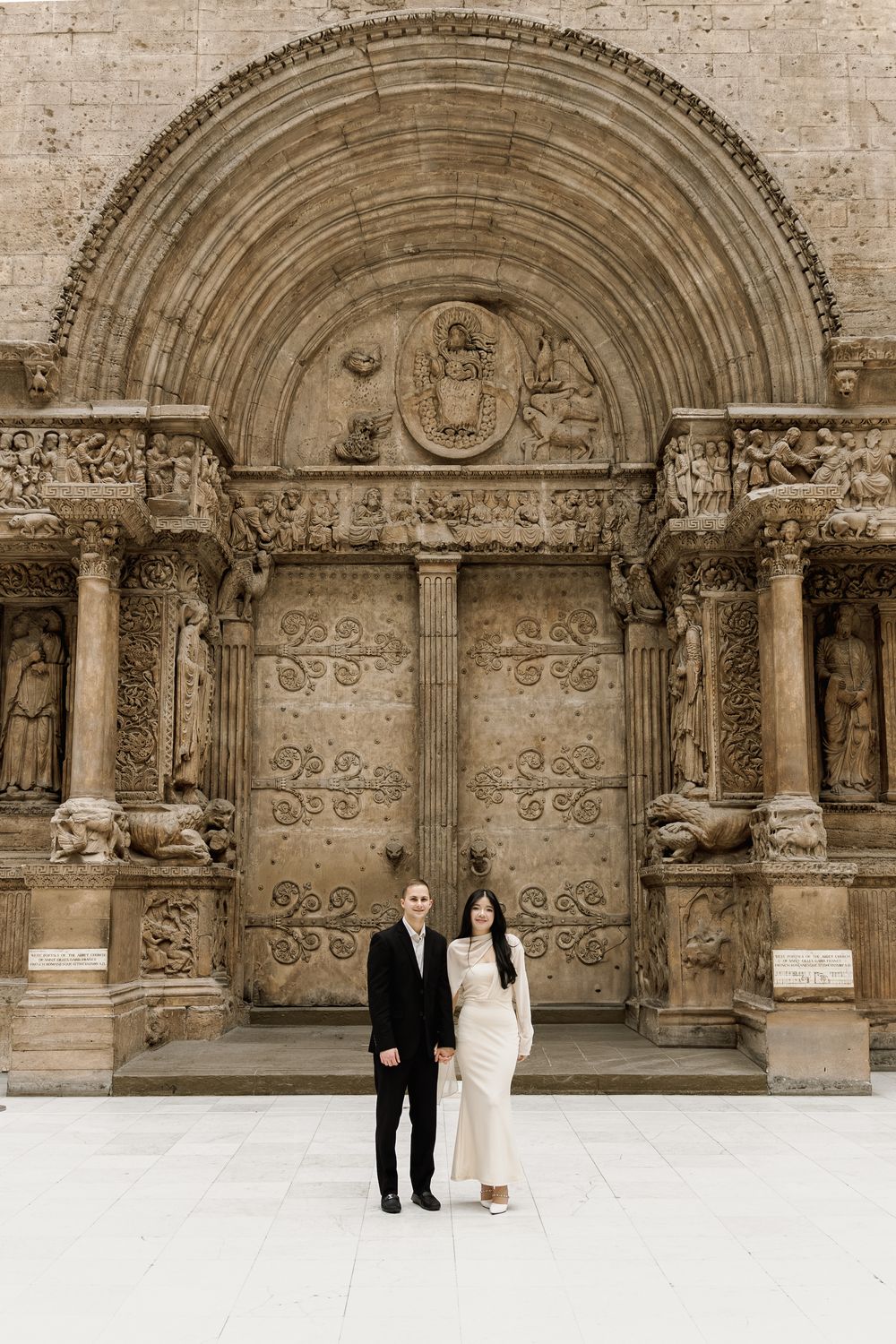 A couple stands in front of an ornate medieval stone church entrance with intricate carved archways and decorative details.