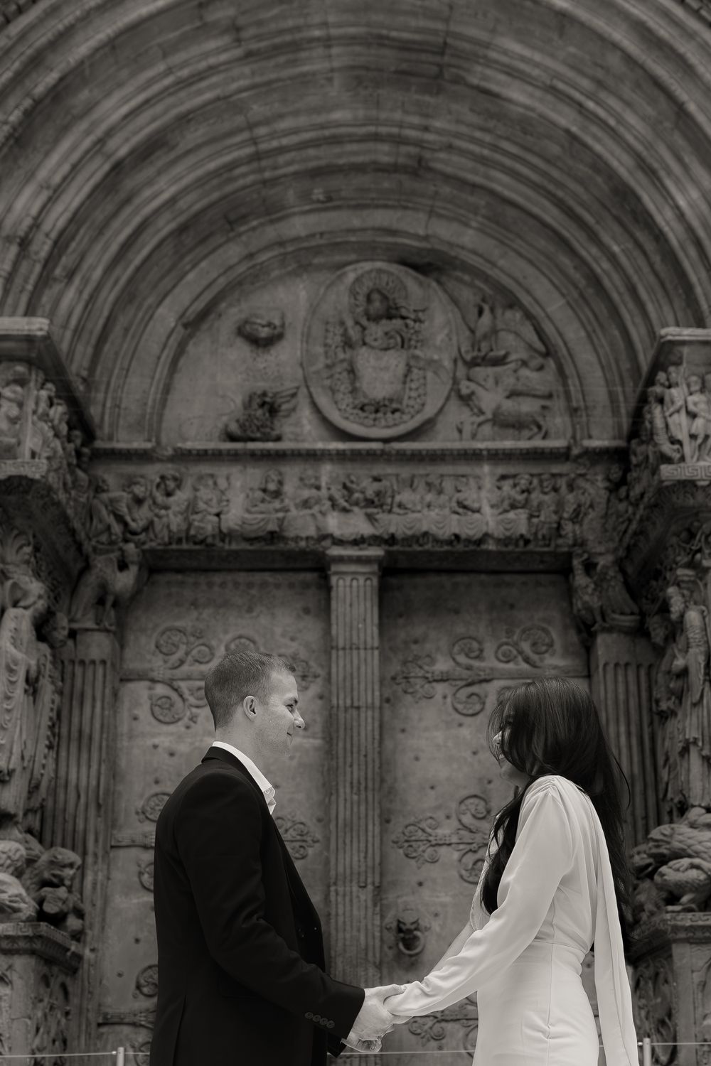Bride and groom stand under ornate stone archway with intricate architectural details in black and white photo.