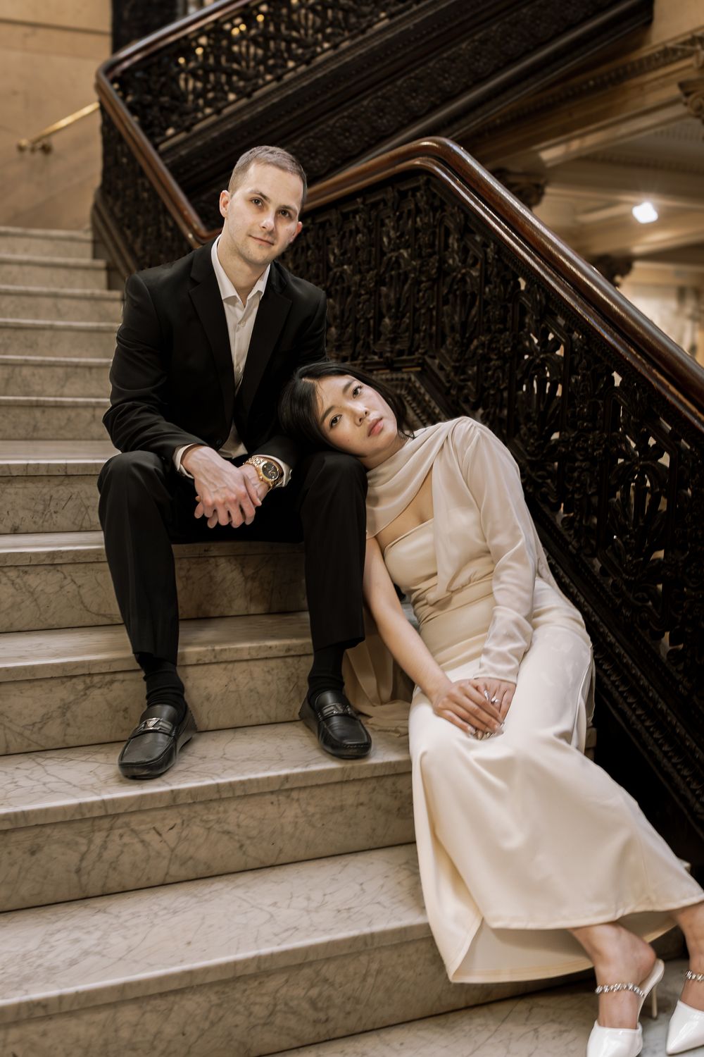 A couple in formal attire poses romantically on elegant stone stairs with an ornate railing.