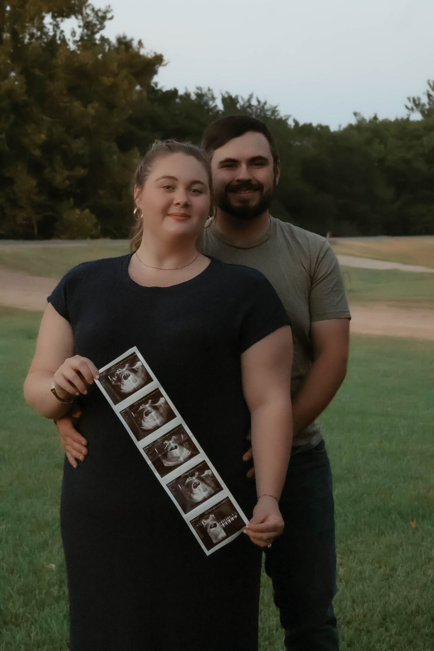 Couple posing together outdoors while displaying ultrasound photos in a grassy field.