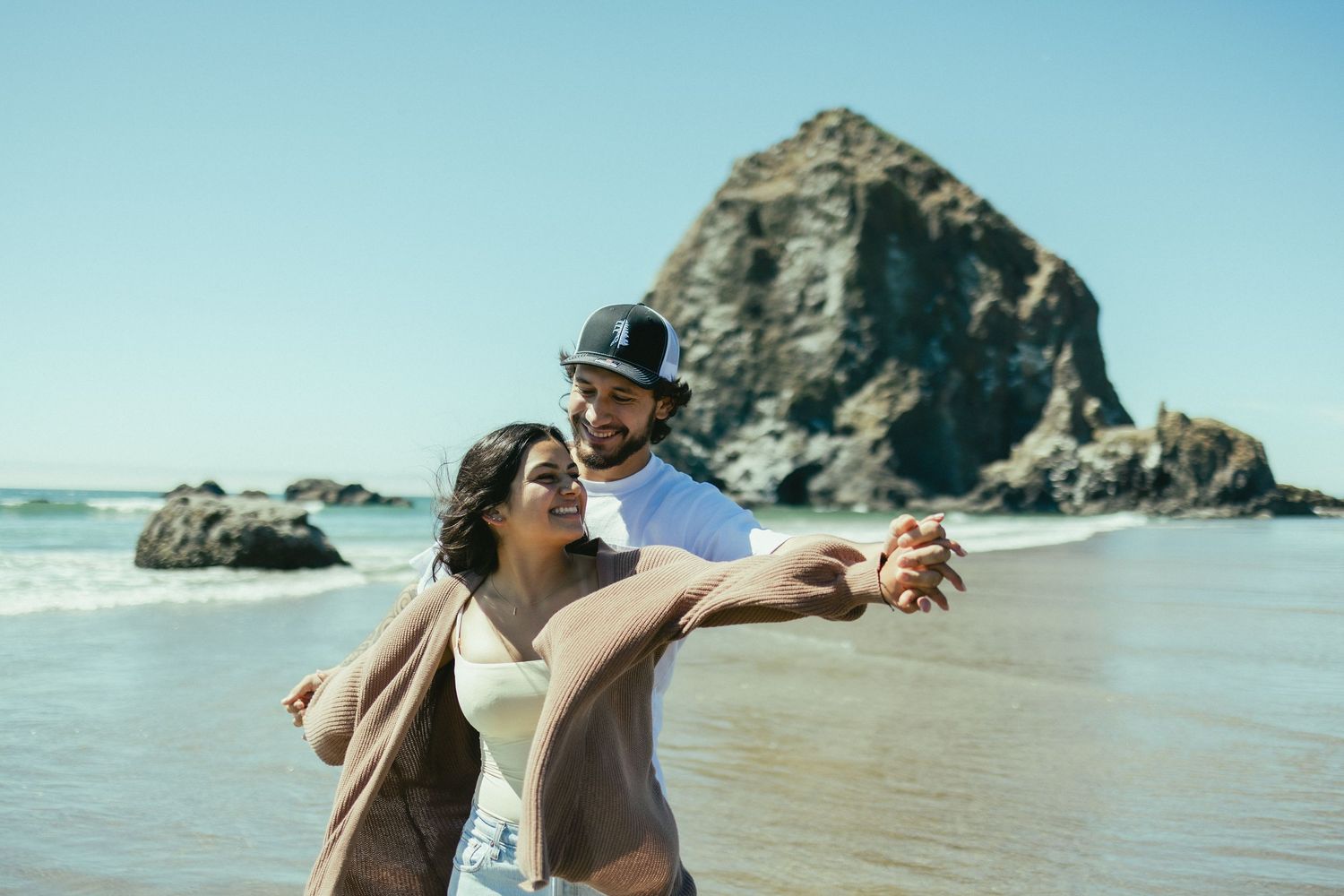 Two people walk along a scenic beach with large rock formations and waves crashing against the shore in Oregon.