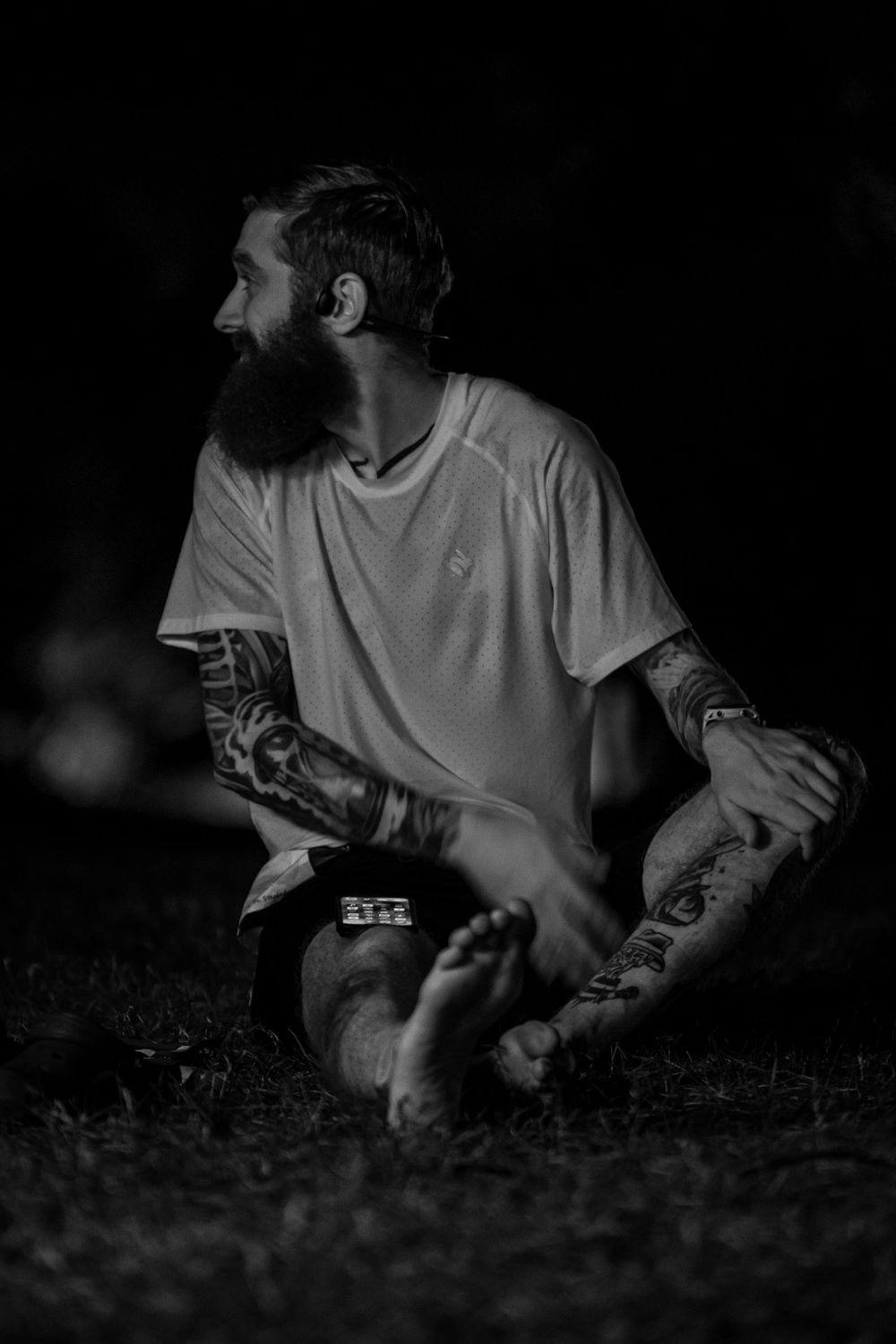 A black and white portrait of a tattooed person sitting on the ground in casual attire.
