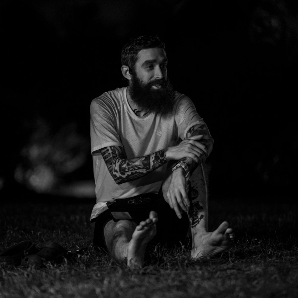 A monochrome image of a bearded person sitting cross-legged outdoors.