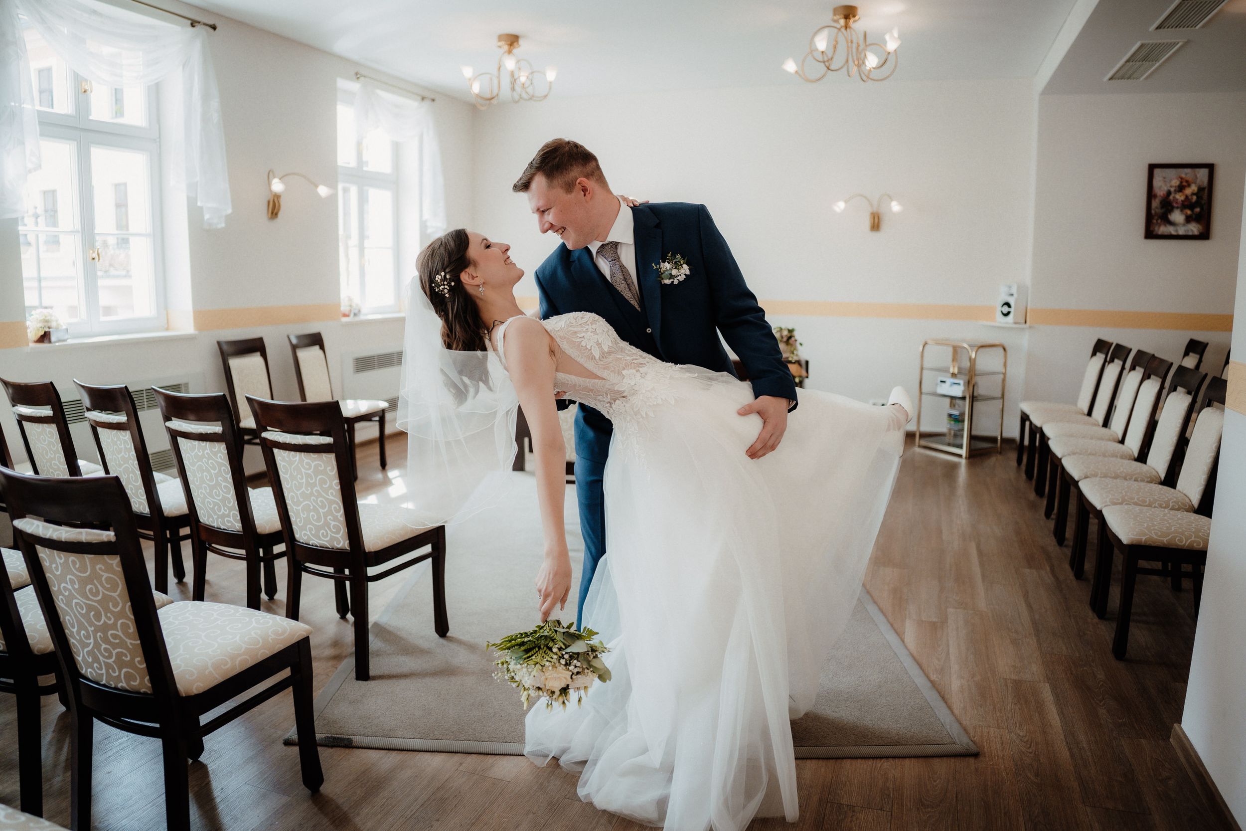 A romantic dip pose during a wedding celebration in an elegant venue with wooden chairs and chandeliers.