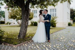 A romantic wedding couple embraces under a tree on a paved walkway near a vintage building.