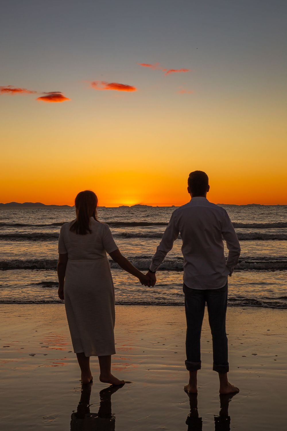 Silhouetted couple holding hands while walking along a beach at sunset with orange sky and ocean waves.