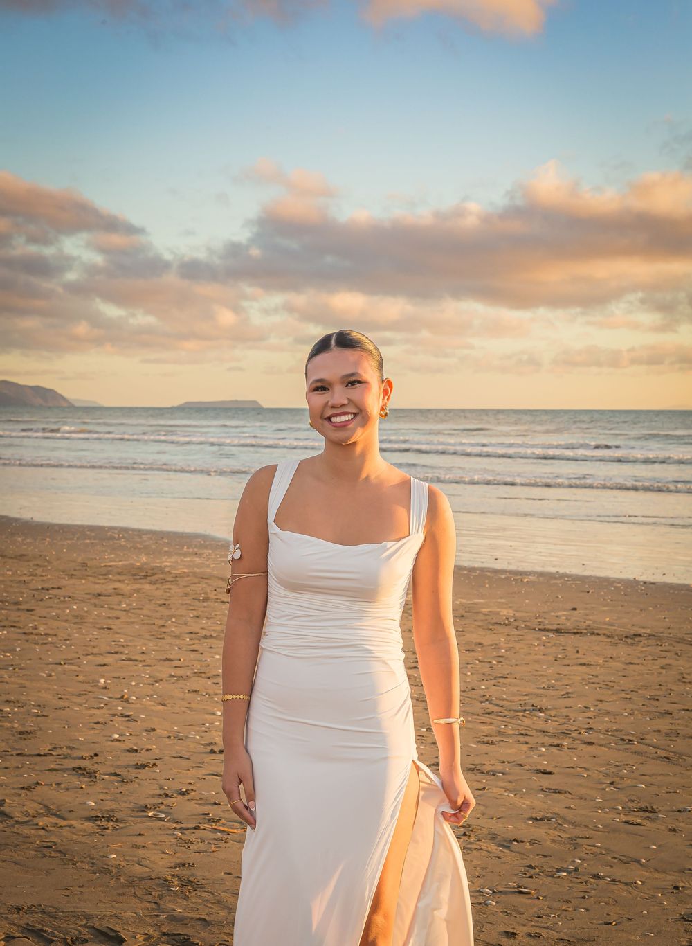 Person in white dress standing on beach at sunset with golden clouds and ocean waves in the background.