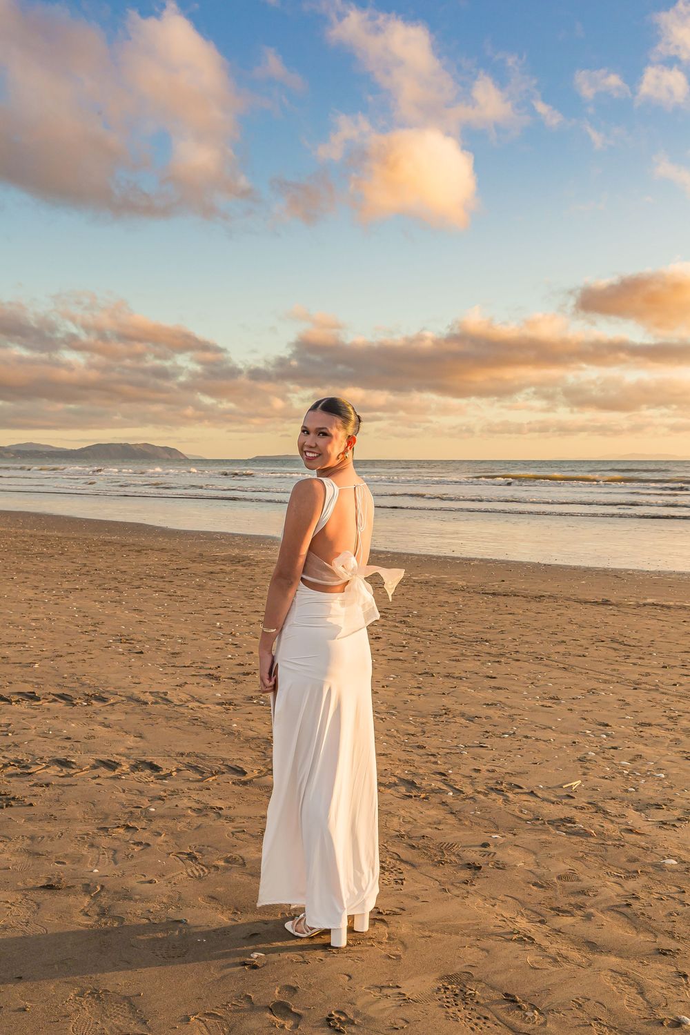 A white formal dress on a beach at sunset with golden clouds and ocean waves in the background.