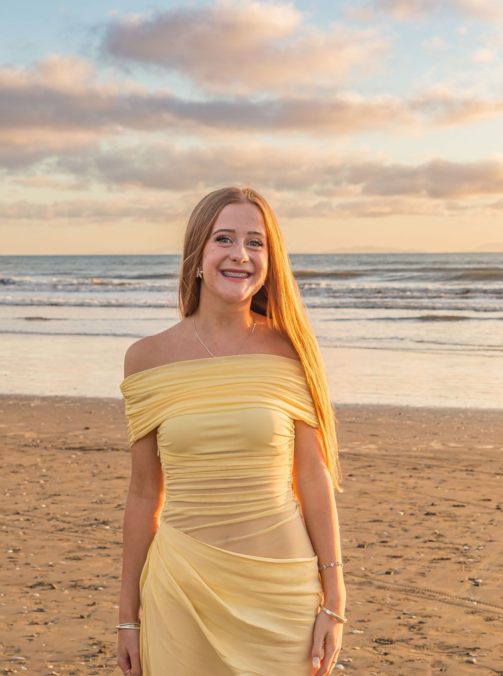 A person in a yellow off-shoulder dress stands on a sandy beach at sunset with ocean waves and cloudy skies in the background.
