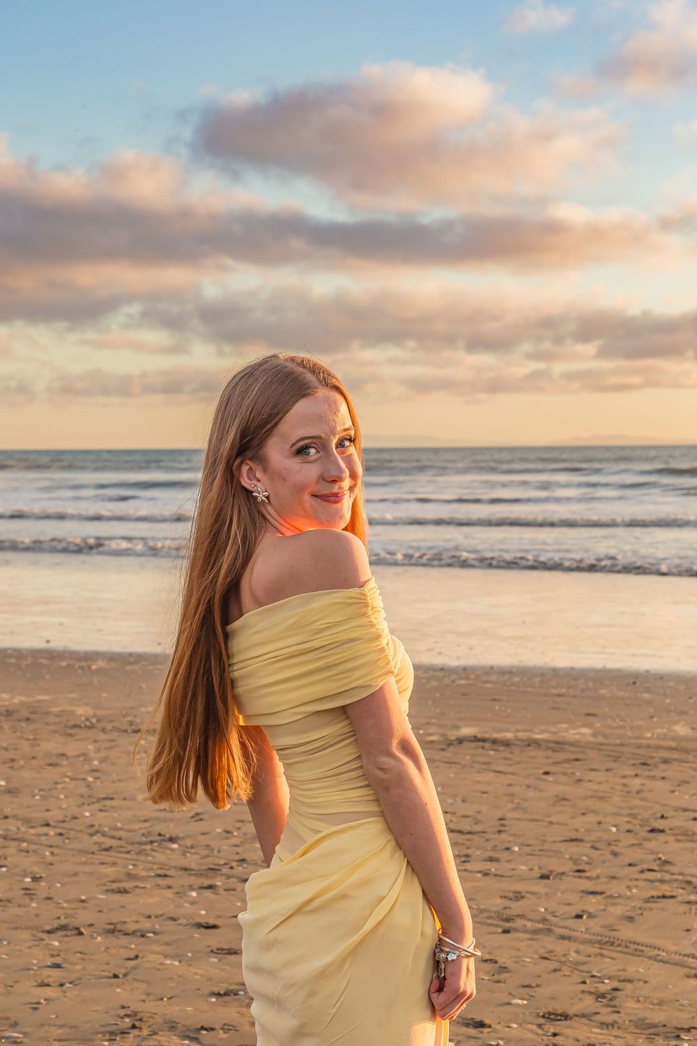 A yellow dress catches the warm sunset light on a beach as waves roll in under pink and orange clouds.