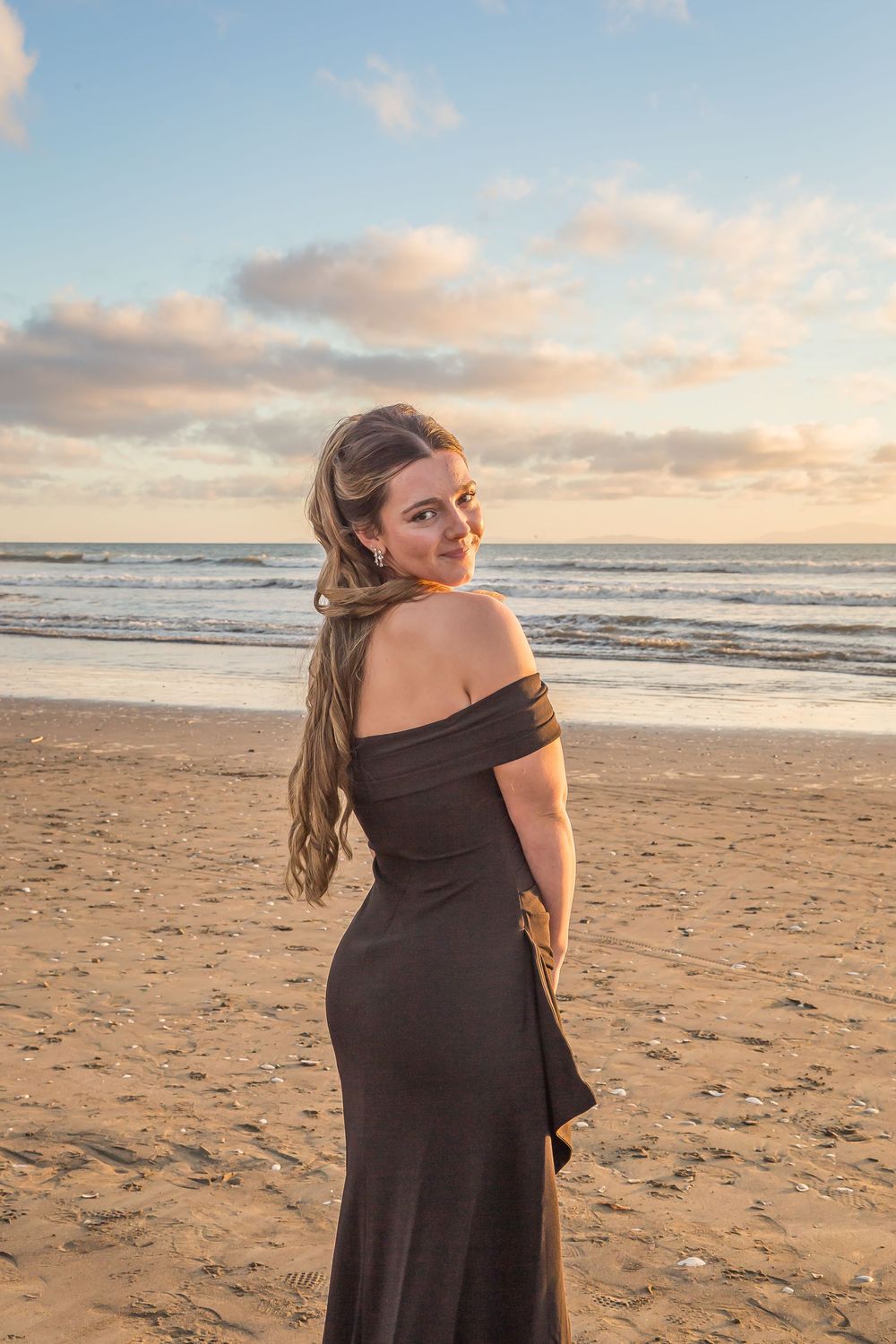 A woman in a dark dress poses on a sandy beach during golden hour sunset with ocean waves in the background.