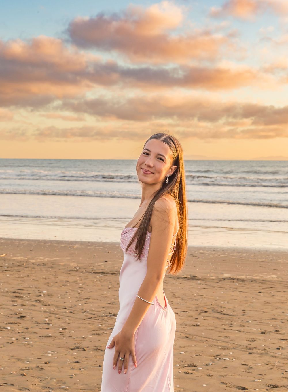 A woman in a flowing pink dress stands on a sandy beach at sunset with orange clouds reflecting on the ocean.
