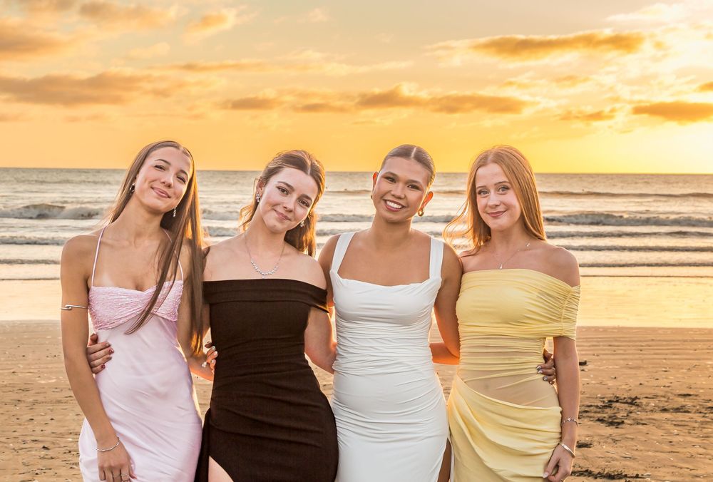 Four friends in elegant dresses pose together on a beach at sunset with golden skies and ocean waves behind them.