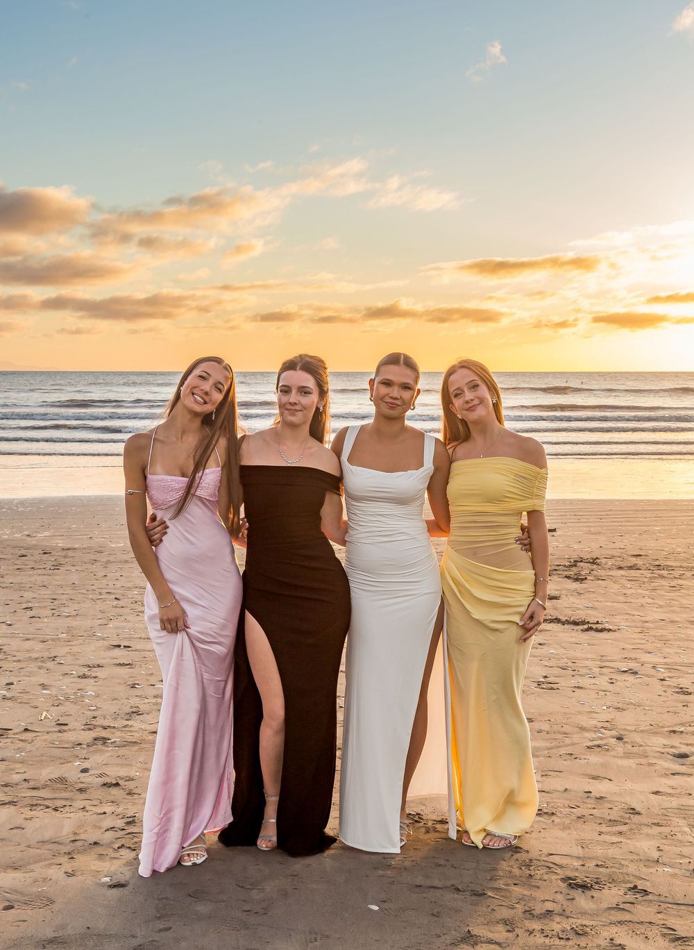Four elegant women in formal evening gowns pose together on a beach at sunset with golden clouds in the sky.