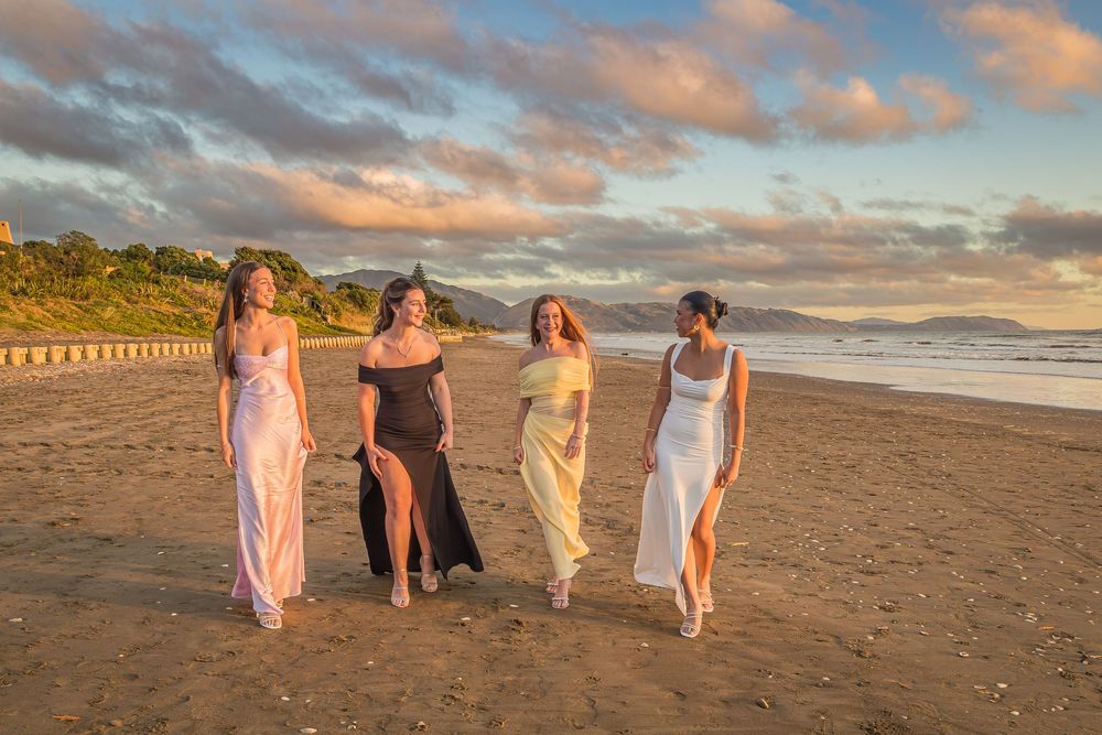 Four women in formal dresses walk along a sunset beach with golden clouds and waves in the background.