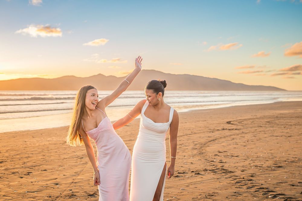 Two people in white dresses enjoying a beautiful sunset together on a sandy beach with mountains in the background.