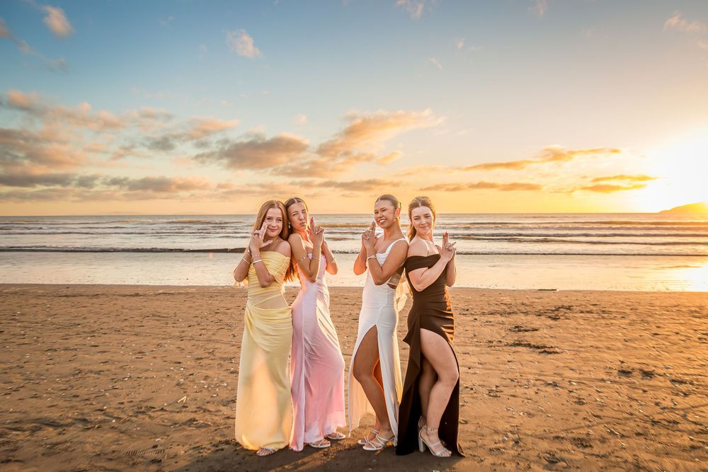 A group poses together on a sandy beach at sunset wearing elegant formal dresses.