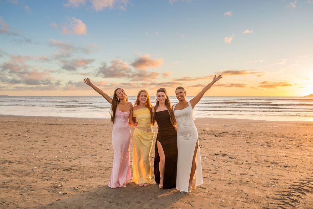 A group in evening gowns poses with raised arms on a beach during sunset with golden clouds in the background.
