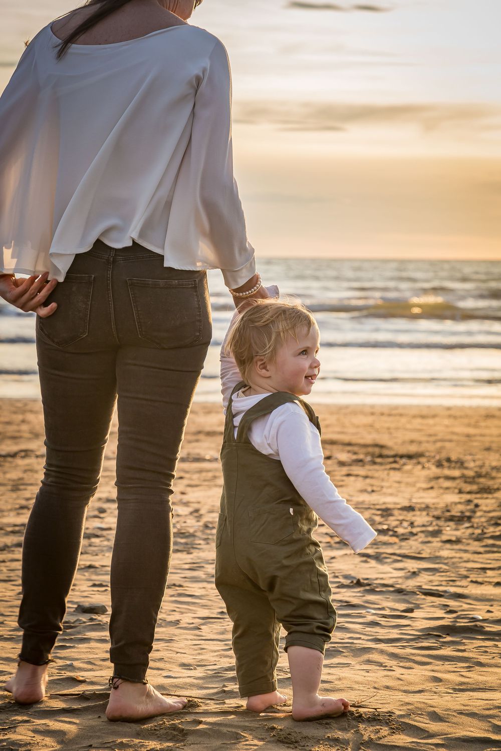 A small child in overalls stands barefoot on a beach at sunset next to an adult, creating a heartwarming silhouette.
