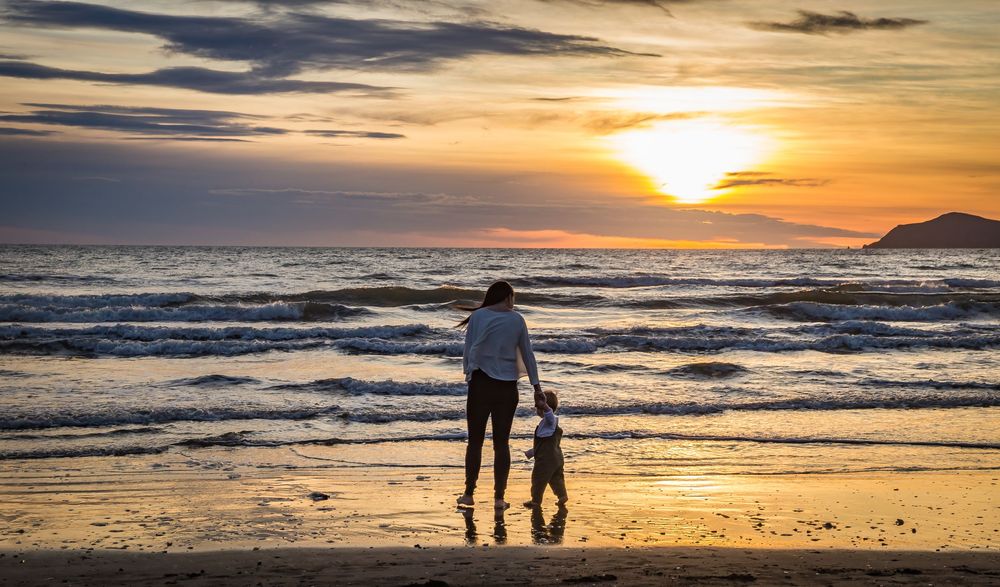Silhouettes of people walking along a beach at sunset with dramatic orange sky and rolling waves in the background.