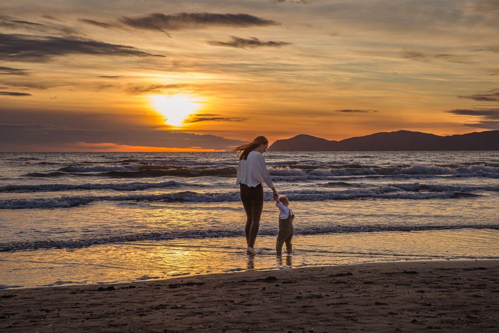 People strolling on a sandy beach during a dramatic orange and yellow sunset over the ocean.