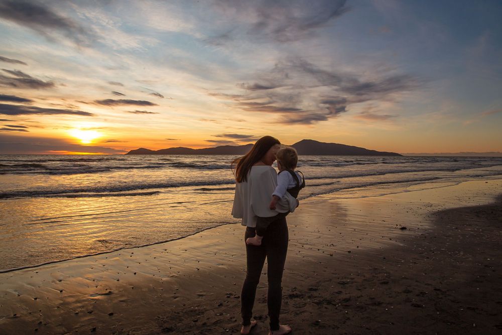 A couple embraces on a sandy beach at sunset with dramatic orange and purple clouds reflecting on the ocean water.