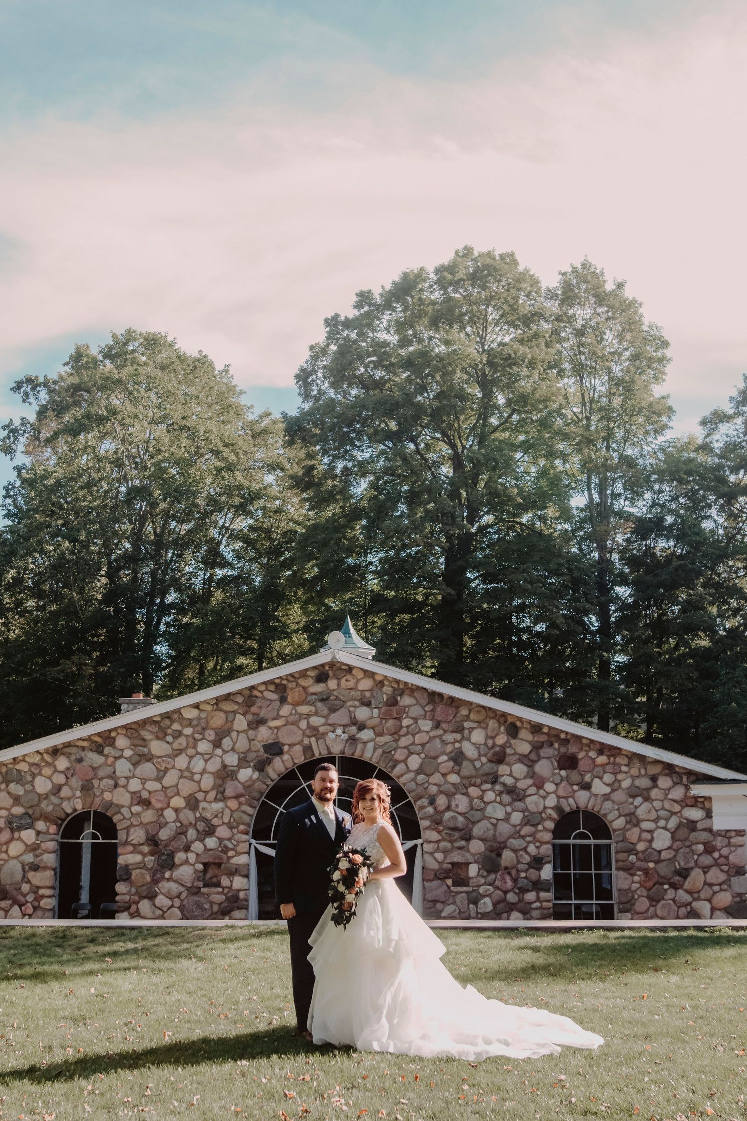 A couple poses in front of a rustic stone chapel surrounded by tall trees.