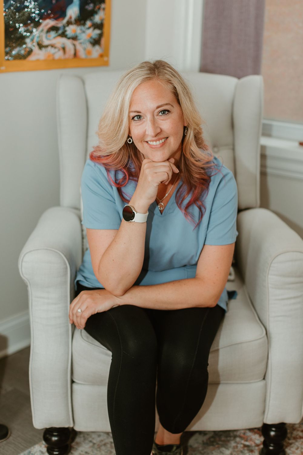 Professional wearing light blue scrubs seated in a grey armchair in an office setting.