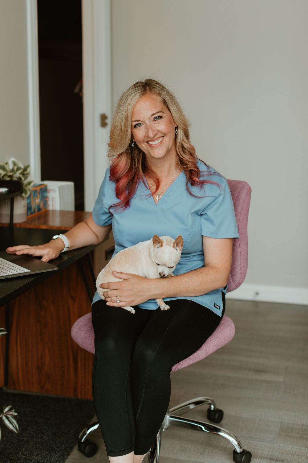 Healthcare professional in blue scrubs sitting in office chair with white cat on lap.