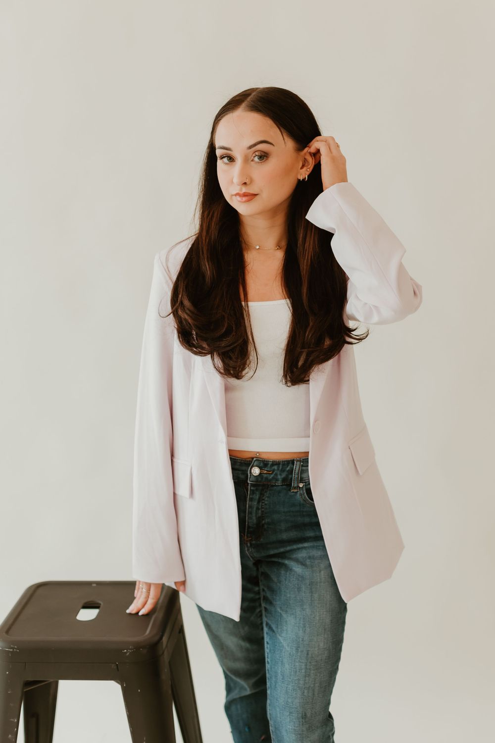 Woman in white blazer and jeans poses casually against light studio backdrop.