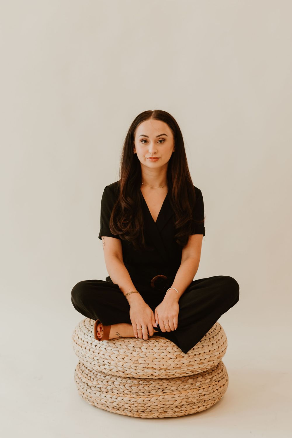 Casual portrait sitting on a woven ottoman pouf wearing all black.