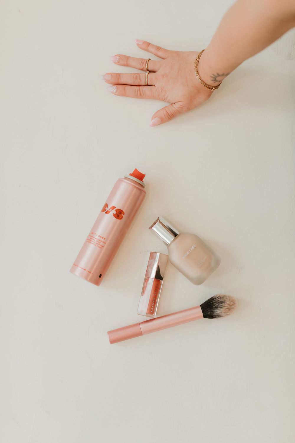 Flatlay of pink makeup products including brush and cosmetics with hand in frame.