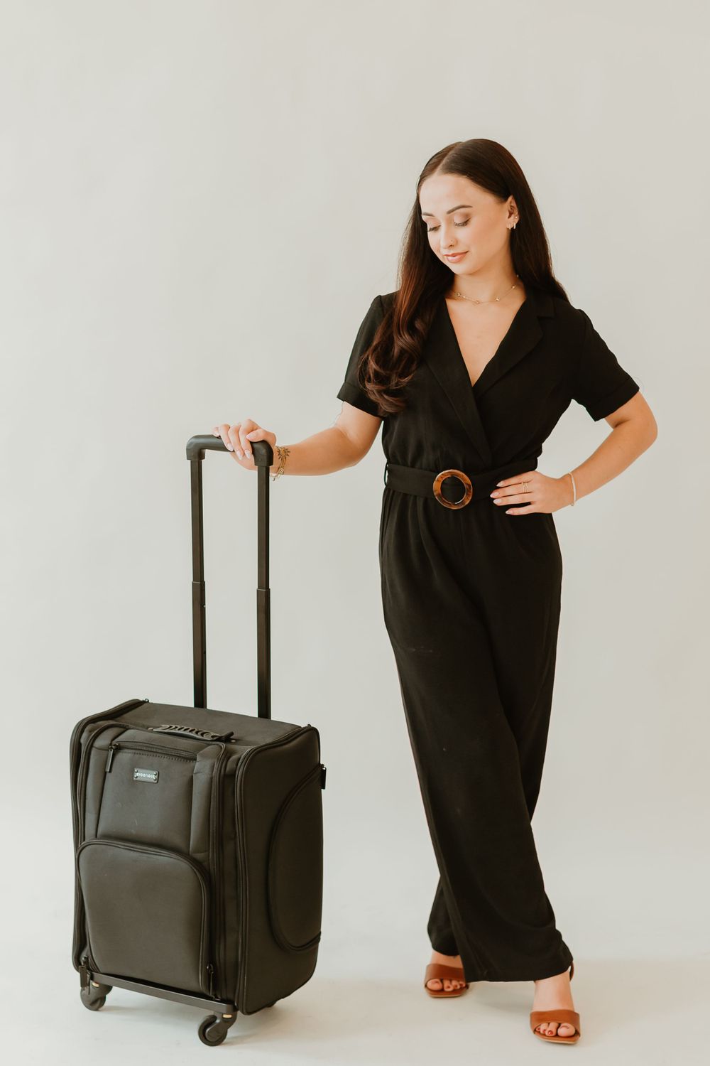 Full length portrait with black jumpsuit and rolling suitcase against white background.