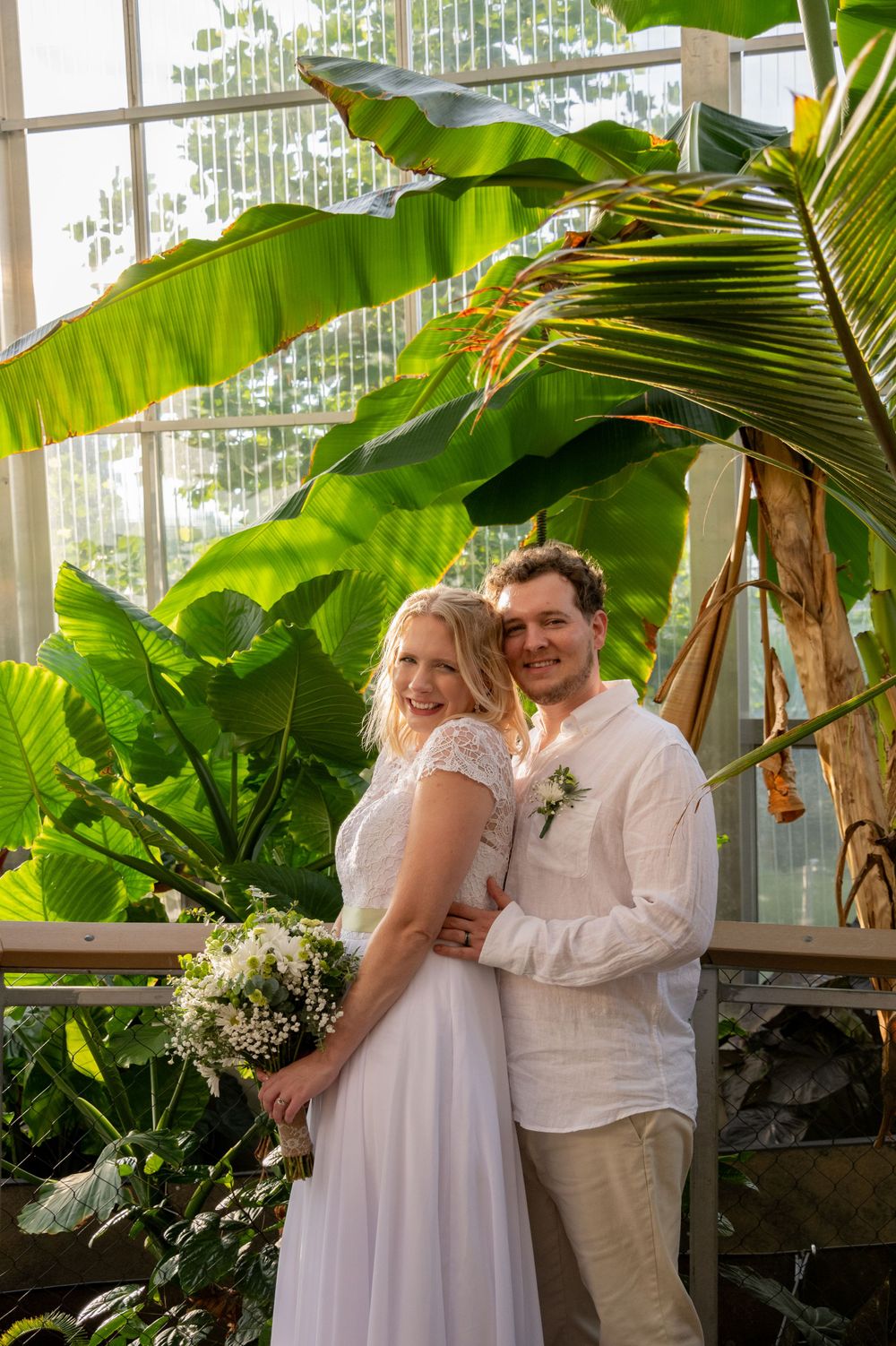 A newlywed couple poses amid lush tropical plants and palm fronds in a greenhouse setting.