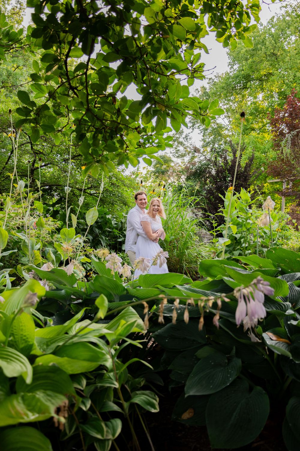 A couple stands embraced in a lush garden pathway surrounded by vibrant green foliage and blooming flowers.