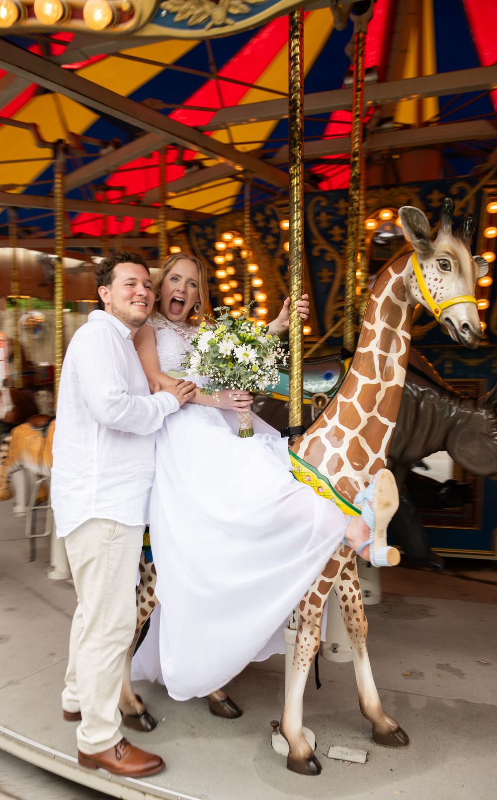 A happy couple rides a carousel giraffe together at an amusement park during their wedding celebration.
