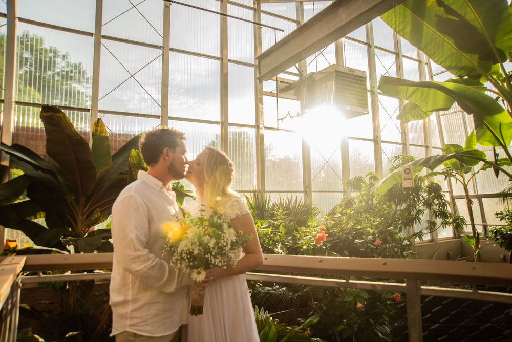 A romantic couple embraces in a sunlit greenhouse filled with lush tropical plants and flowers.
