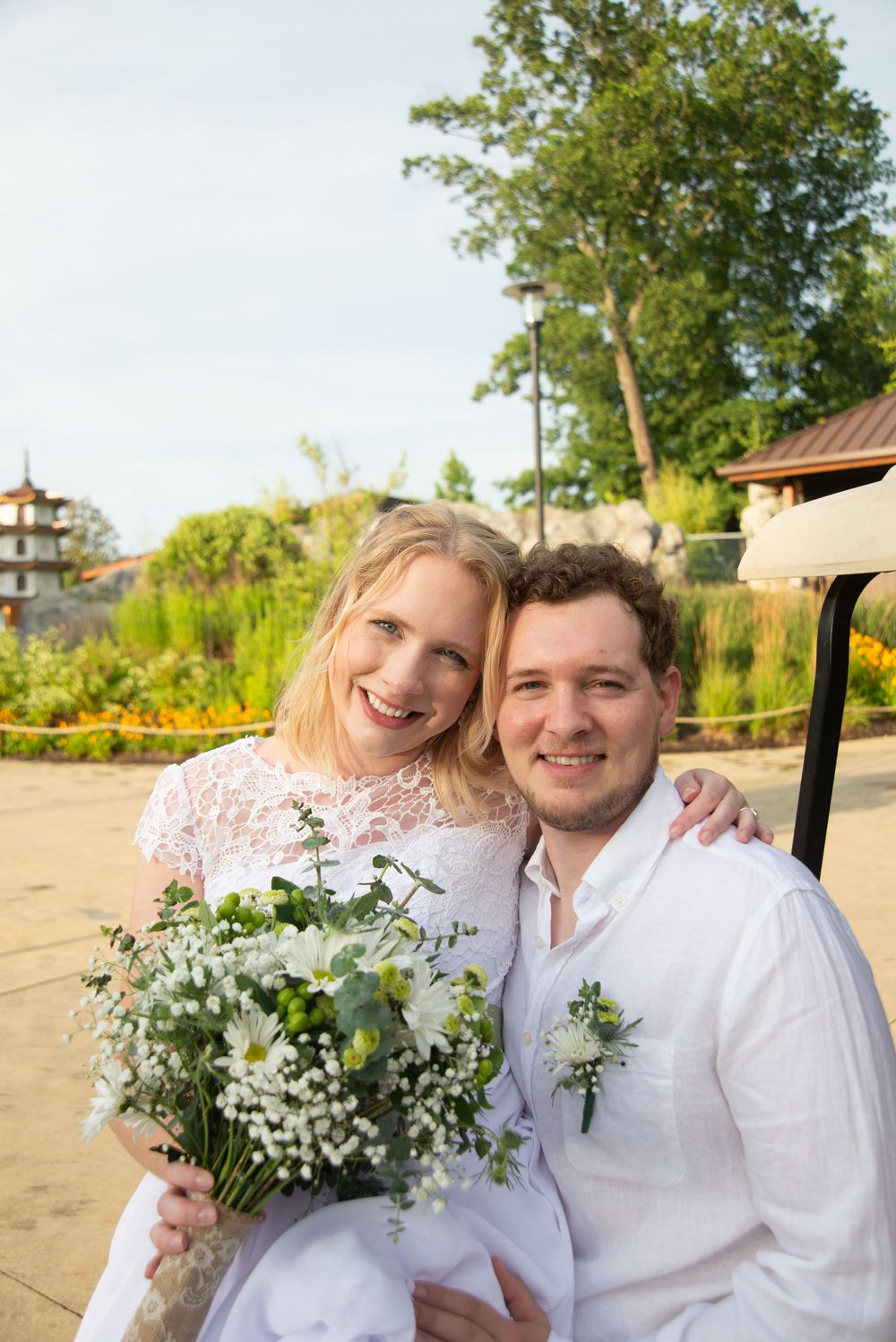 A couple in wedding attire sits together outdoors holding a delicate bouquet of white and green flowers.
