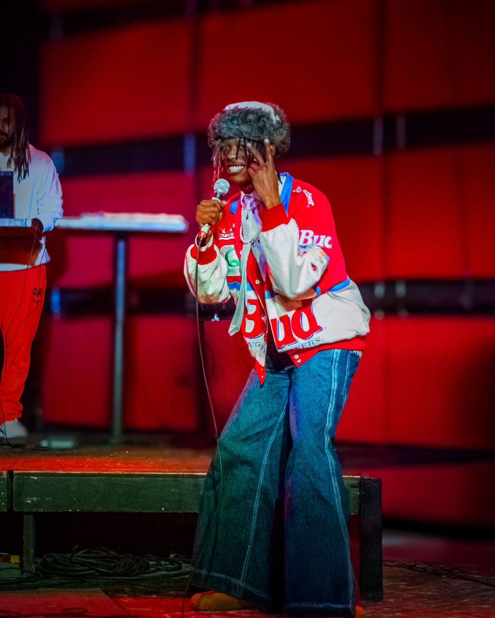 A performer in denim overalls sings passionately into a microphone against a dramatic red-lit backdrop.