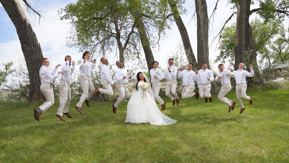 Bride in white gown surrounded by groomsmen in tan suits jumping joyfully on green lawn with trees backdrop.