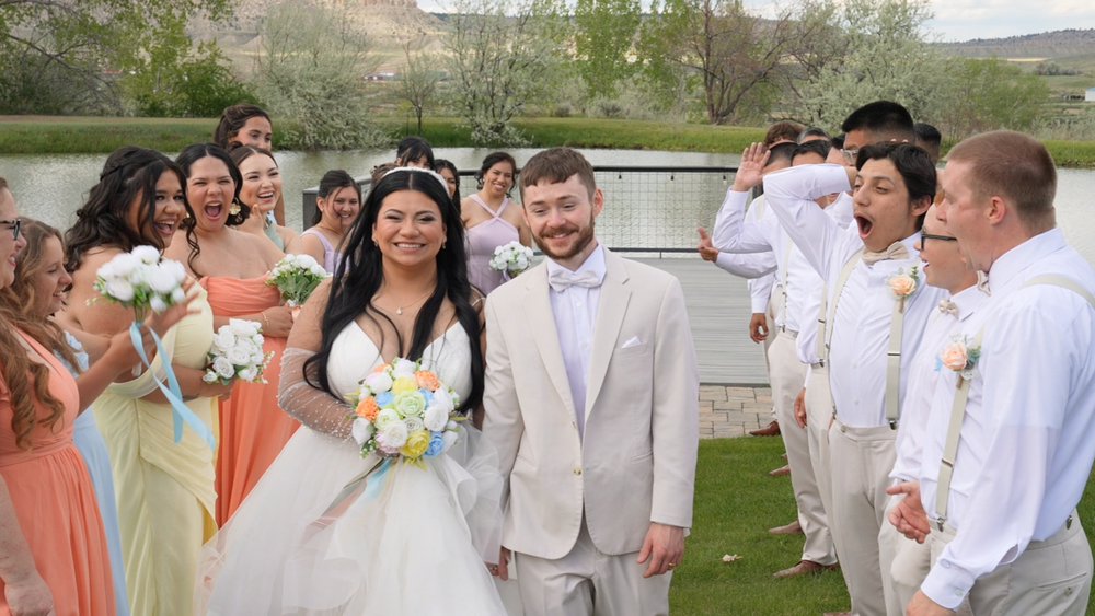 Bride and groom walk joyfully through cheering wedding party outdoors, bridesmaids in peach and yellow dresses holding bouquets.