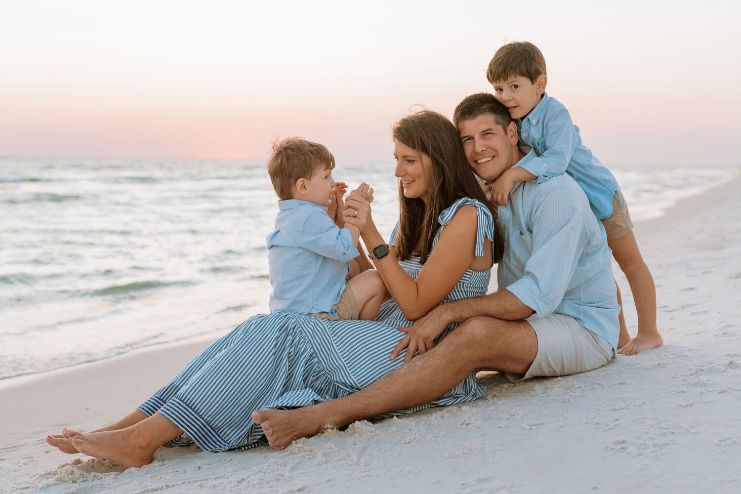 A family wearing coordinating blue outfits sits together on a white sandy beach at sunset.