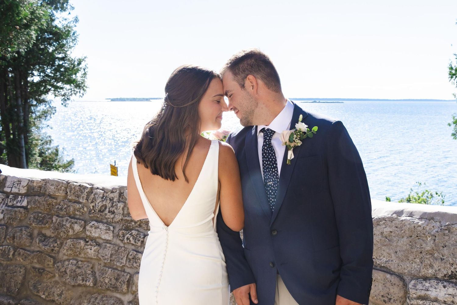 Romantic couple embraces by stone wall overlooking ocean on sunny wedding day in coastal setting.