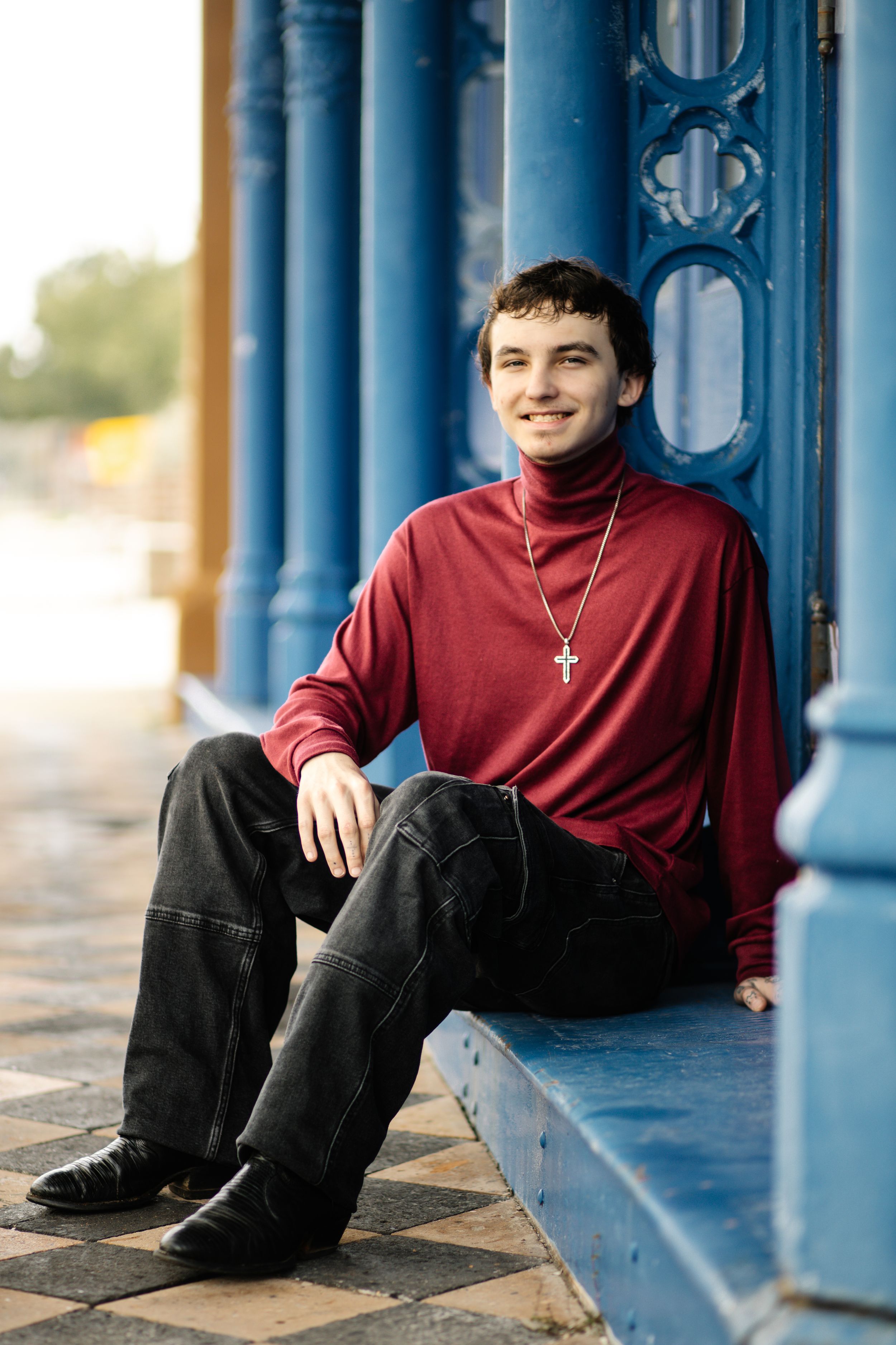 Young person in burgundy turtleneck and dark pants sits casually against blue architectural columns outdoors.