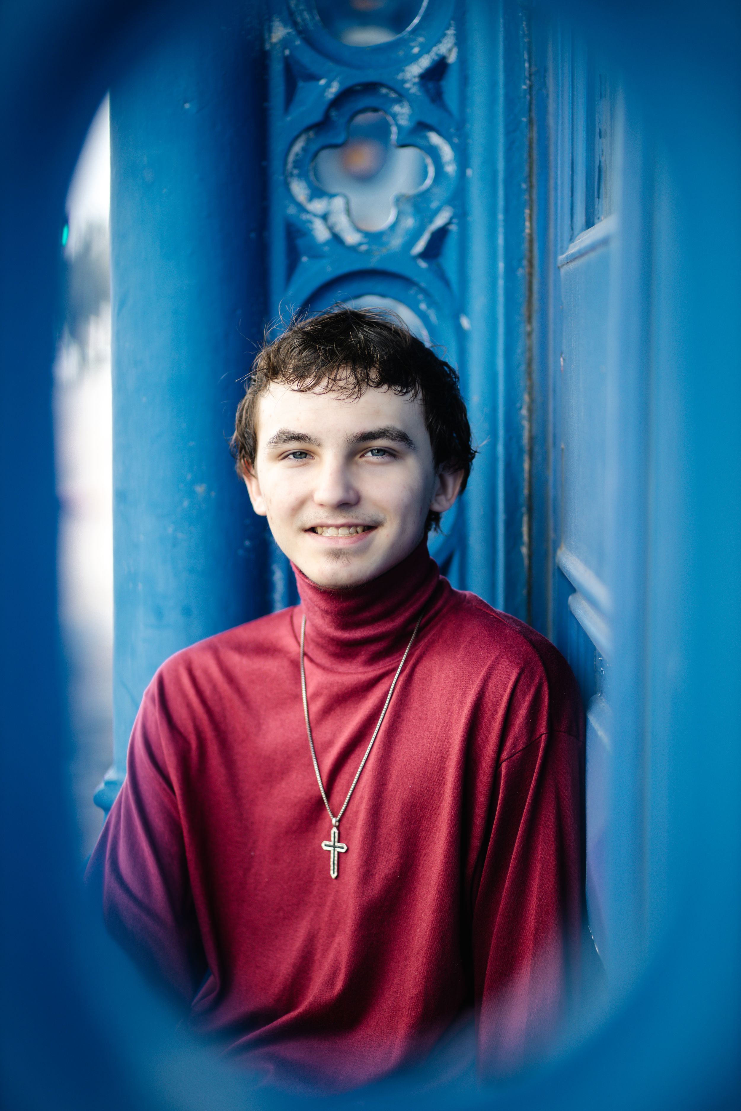A figure in a red turtleneck sweater with a cross necklace poses against an ornate bright blue background.