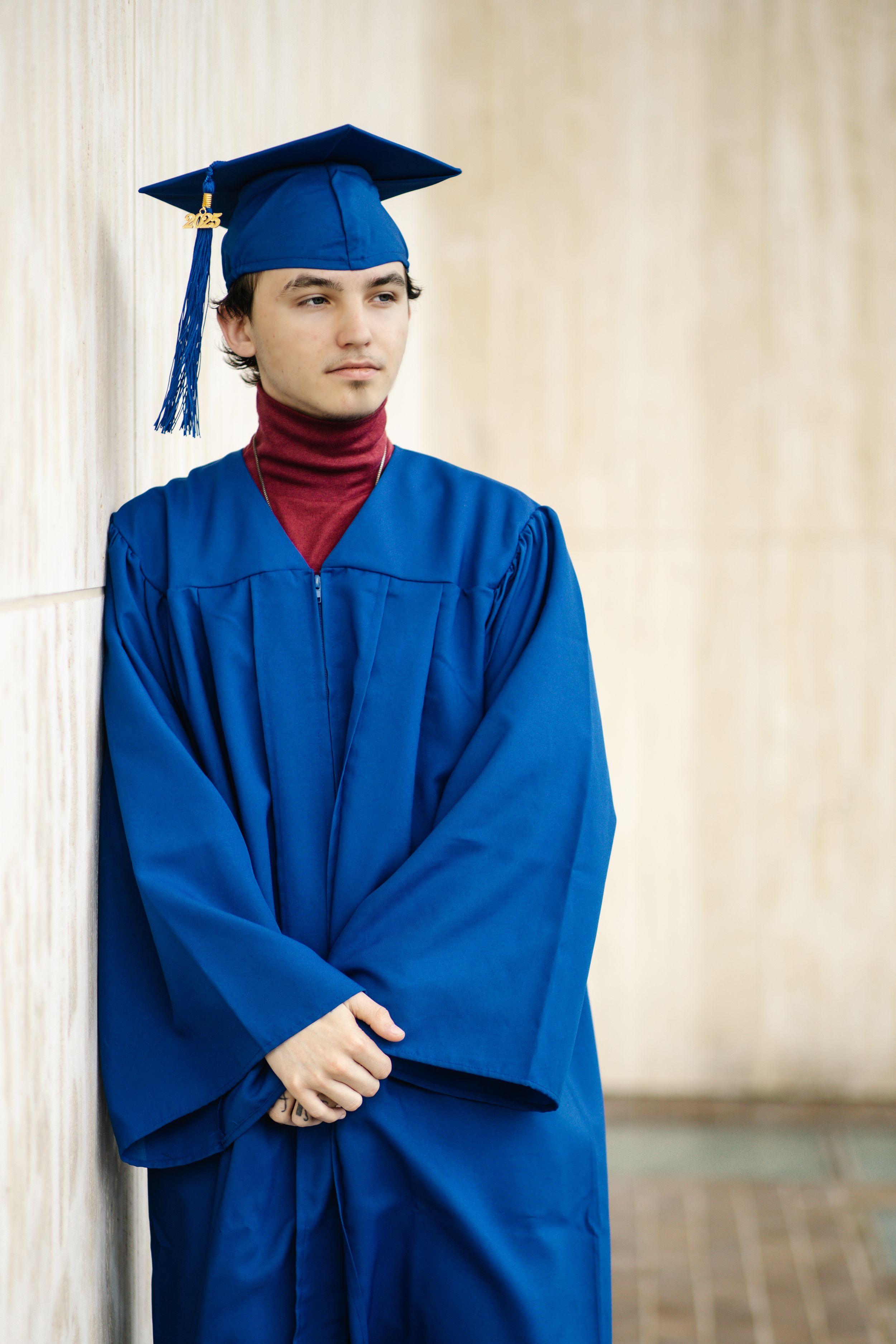 Graduate wearing blue cap and gown with red turtleneck stands against beige wall during commencement ceremony.