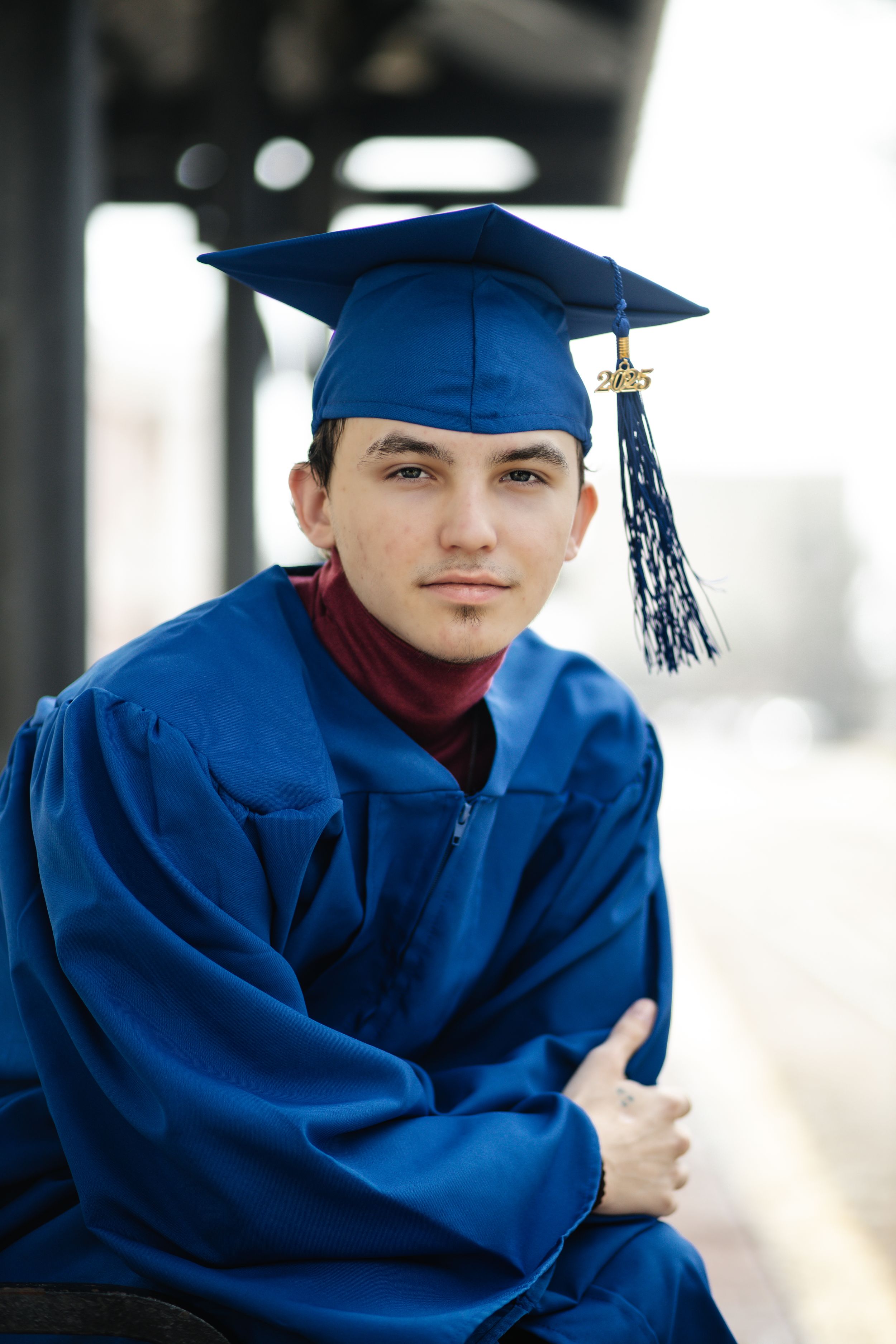 A graduate wearing a royal blue cap and gown with a burgundy turtleneck sits for a portrait.